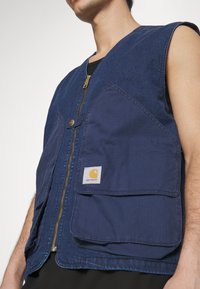 Granatowa kamizelka z wytrzymałego materiału, z zamkiem błyskawicznym z przodu, dwiema dużymi kieszeniami z przodu oraz wyraźnym logo Carhartt na lewej kieszeni.