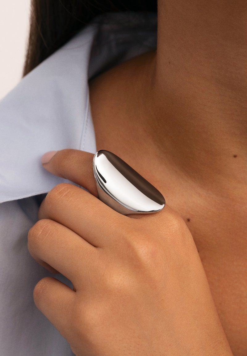 Anillo de metal con una superficie pulida y lisa y una parte superior redondeada. Lleva en el dedo índice, complementado con un cuello de camisa de color azul claro.