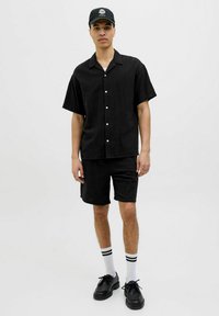 Camisa negra de manga corta con botones en el frente y textura lisa, combinada con pantalones cortos a juego. Zapatos negros y calcetas hasta el tobillo con rayas blancas y negras.