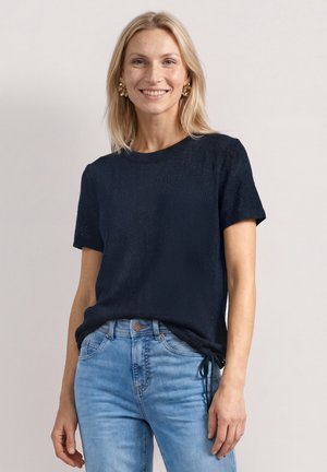 Smilende kvinde med blondt hår iført sort kortærmet top og lyseblå jeans, stående mod en ensfarvet lys baggrund.