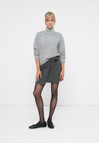 Grå strikket sweater med høj hals, parret med en ternet omslagsskjørt med sidelukning. Person iført gennemsigtige sorte strømpebukser og flade sko.