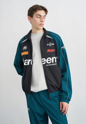 Noor mees, kes kannab türkiissinist ja musta motorspordijopet ning vastavates värvides pükse, vaatab alla ja kõrvale, taust on ühevärviline.