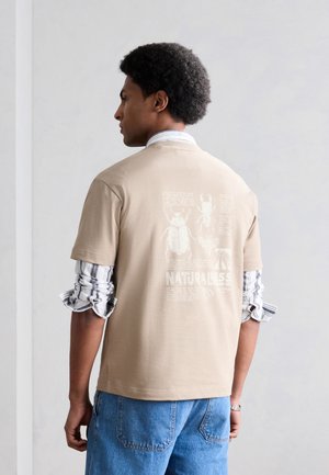 Άνδρας με σηκωμένα ριγωτά μανίκια πουκάμισου φοράει μπεζ μπλουζάκι με ζωγραφιές έντομα και το κείμενο «NATURALISS» στην πλάτη, σε συνδυασμό με μπλε τζιν.