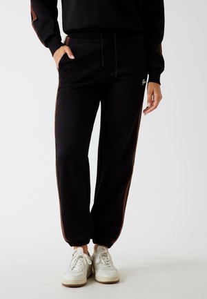Pantaloni jogger neri con strisce laterali marroni, vita elasticizzata e polsini, dotati di una tasca laterale e una texture in tessuto liscia e morbida.