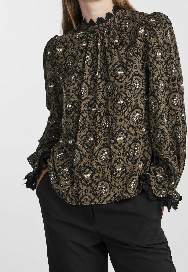 Femme portant un chemisier à manches longues marron à motifs avec une bordure en dentelle noire au col et aux poignets, associée à un pantalon noir.