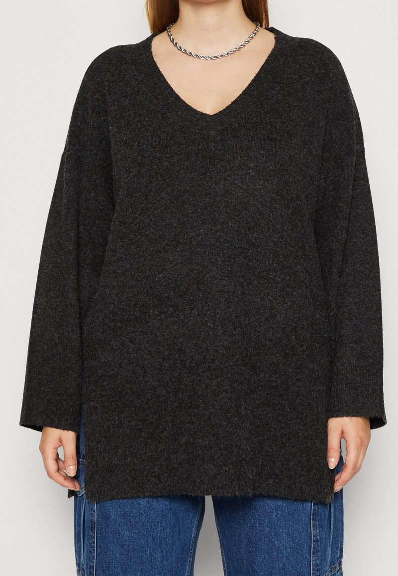 Personne portant un pull ample gris foncé à col en V et un pantalon en denim bleu, accessoirisé d'un collier en chaîne torsadée argentée.