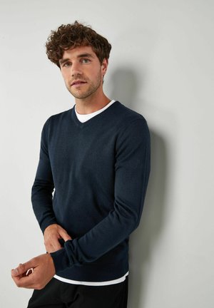 Marineblå v-hals sweater i blødt stof, tætsiddende design, lange ærmer, ribbede manchetter, båret over en hvid rundhalset skjorte, model foran en grå væg.
