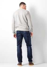 Mand vendt mod væg iført en lysegrå sweatshirt, mørkeblå jeans og brune sko, stående med hænderne langs siden.