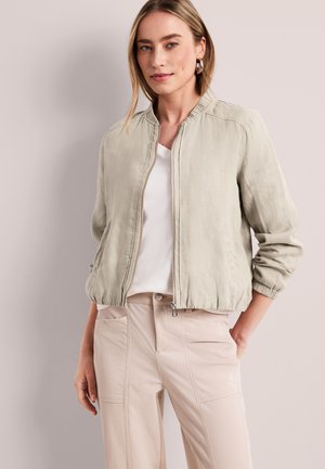 Kvinde iført en lysebeige bomberjakke over en hvid skjorte og beige bukser, stående foran en ensfarvet bleg baggrund.