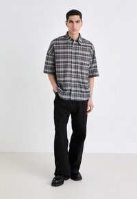 Camicia a maniche corte a quadri in bianco e nero con bottoni davanti, abbinata a pantaloni neri larghi e scarpe nere chunky. Sfondo semplice.