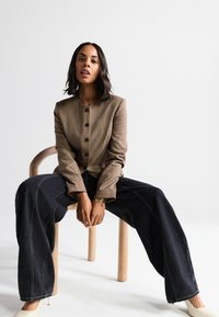 Sacou maro cu model pătrat, guler rotund și nasturi negri, asortat cu blugi largi din denim închis și pantofi de culoare crem.