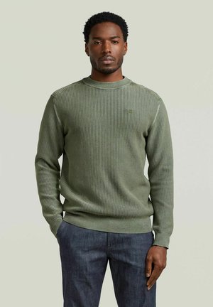 Uomo che indossa un maglione verde oliva con trama e jeans scuri, in piedi con una mano in tasca davanti a uno sfondo semplice.