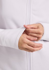 Giacca bianca con zip realizzata in un morbido e liscio tessuto. In primo piano si vedono mani che afferrano il polsino e la zip, mettendo in risalto i dettagli delle cuciture e la texture del materiale.