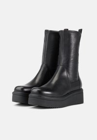 Botines de cuero negro con una textura suave, puntera redondeada y suela gruesa de plataforma. Presenta paneles laterales elásticos y lengüetas para facilitar el calzado.