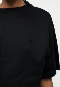 Personne portant un t-shirt noir uni à manches courtes avec un petit logo brodé discret sur le côté gauche de la poitrine.