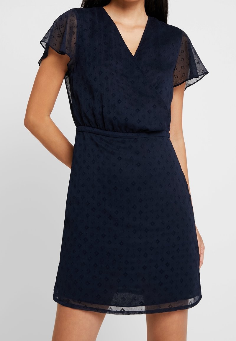 Femme portant une robe bleu marine à manches courtes, col en V, manches volantées et motif subtil à pois, debout avec un bras dans le dos.