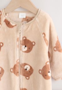 Body bébé beige doux avec des visages d'ours en peluche marron imprimés, avec une fermeture éclair devant et un col rond, suspendu sur un cintre en bois.