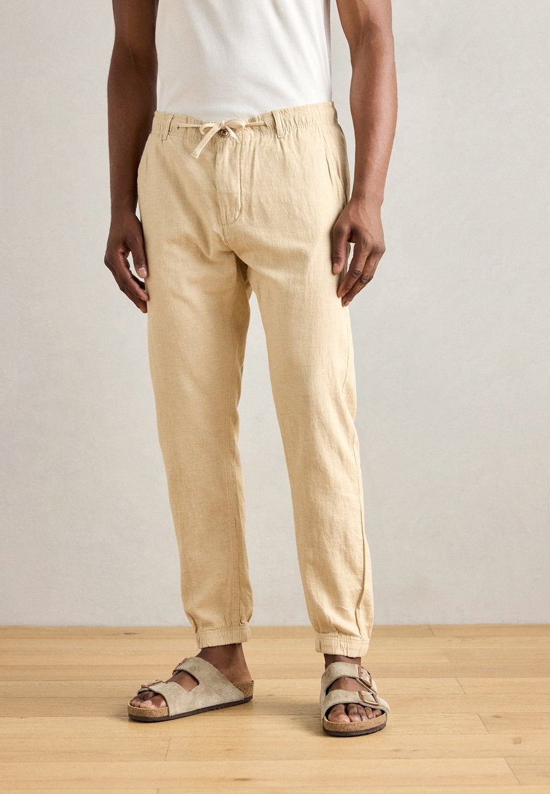 Beige linnen broek met een elastische tailleband en trekkoord, met een ontspannen pasvorm en opgerolde zomen, gecombineerd met open teen grijze sandaal.