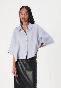 Camicia button-up oversize grigia chiara con colletto, maniche corte e vestibilità rilassata, abbinata a una gonna nera in pelle.