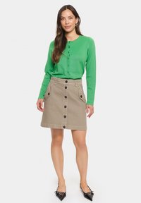 Top in maglia verde a maniche lunghe con bottoni, abbinato a una gonna di jeans beige, con tasche frontali e bottoni neri al centro. Scarpe nere a punta.