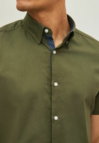 Camisa de manga corta verde oliva con cuello abotonado. Cuello interior con patrón azul. Textura suave con botones blancos.
