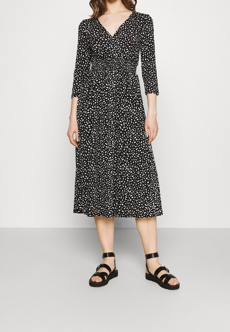 Czarna sukienka kopertowa z białym abstrakcyjnym wzorem, z rękawami 3/4, marszczonym w pasie i długości midi; połączona z czarnymi sandałami na paski.