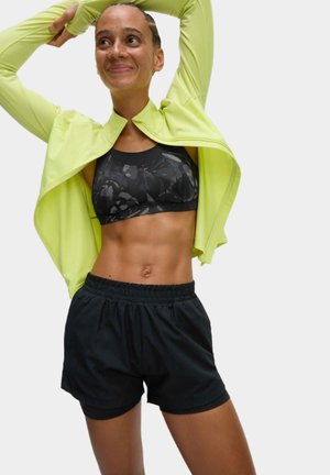 Geel lichtgewicht jas over een zwart gepatineerde sport-bh, gecombineerd met zwarte sportshorts met elastische taille. Gladde texturen en een aansluitend ontwerp.
