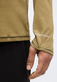 Κοντινό πλάνο χειρός εν μέρει καλυμμένης από μία μπεζ, υφή μακριάς μανίκας με άνοιγμα για τον αντίχειρα και το κείμενο "LIGHTRANGE" στην περιοχή του καρπού.