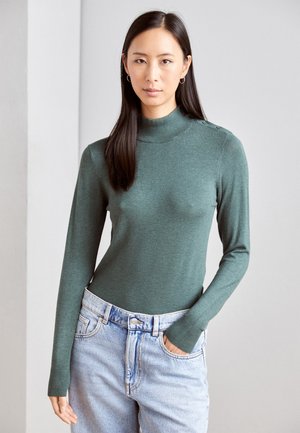 Pull en col roulé à manches longues vert en tissu doux, avec des poignets côtelés et des détails de boutons aux épaules, associé à un jean en denim clair.