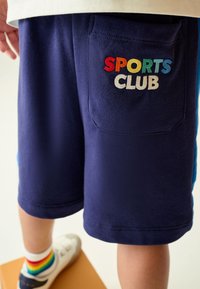 Tamsiai mėlynos šortai iš minkštos medžiagos su užpakaline kišene, kurioje yra spalvotu siuvinėjimu užrašytas tekstas „SPORTS CLUB“. Šortai turi laisvo kirpimo siluetą.