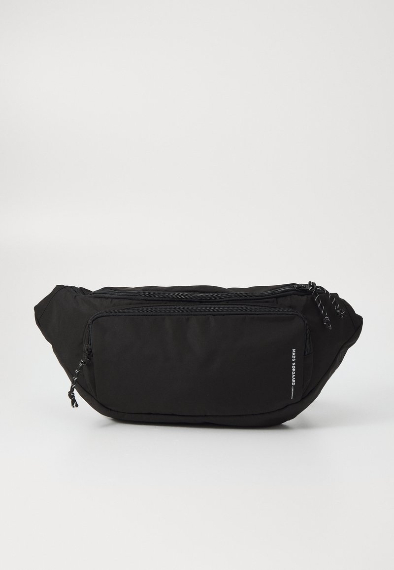 Banane en tissu noire avec poche zippée à l’avant et texte de marque sur le devant, présentée sur un fond clair uni.
