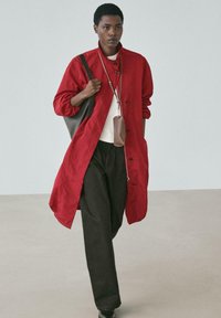 Roter Leinen-Langmantel mit Knöpfen, kombiniert mit einem weißen Oberteil und dunklen Hosen. Accessoires umfassen eine braune Umhängetasche, die an einem Riemen hängt.