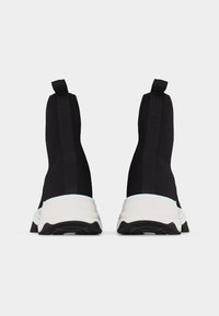 Ζευγάρι μαύρα αθλητικά παπούτσια τύπου κάλτσας με λευκές χοντρές σόλες, από πίσω, σε απλό ανοιχτό γκρι φόντο.