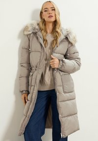 Abrigo ligero de plumas beige con capucha forrada de piel, cintura ajustada, textura acolchada y detalles de botones a presión, combinado con un suéter de punto trenzado.