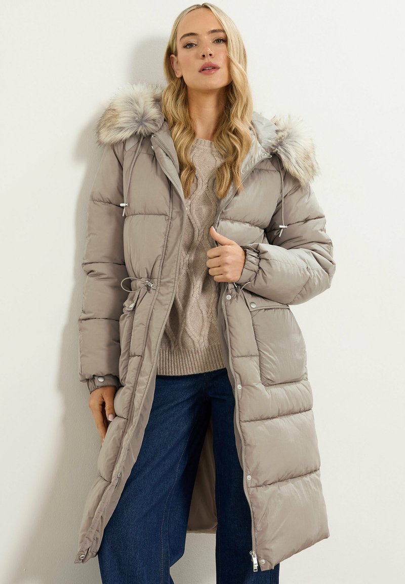 Abrigo ligero de plumas beige con capucha forrada de piel, cintura ajustada, textura acolchada y detalles de botones a presión, combinado con un suéter de punto trenzado.