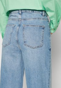 Jeans de denim azul claro con cintura alta, dos bolsillos traseros y un diseño de pierna recta. La textura es suave con un ligero desvanecimiento.