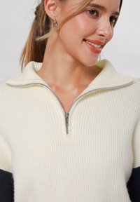 Maglione a coste crema con colletto zip, maniche in blu scuro e una texture morbida. I dettagli della cerniera argento aggiungono un contrasto.