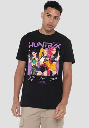 Young man wearing black T-shirt with colorful illustration of three animated female K-pop singers holding microphones and stylized text "HUNTRX."