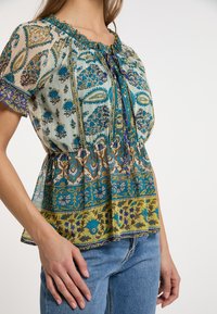 Blusa leggera con stampa floreale, colletto arricciato, maniche a palloncino e vita a peplo. Presenta motivi intricati in blu, verde e giallo.