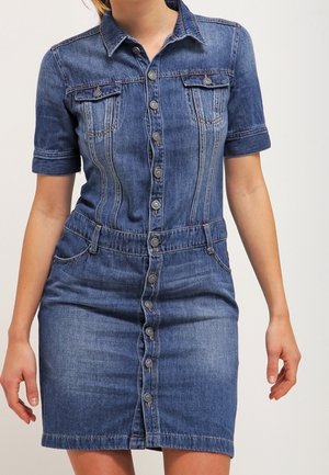 Kvinde iført en kortærmet denim kjole med knapper foran, krave og forlommer mod en ensfarvet baggrund.