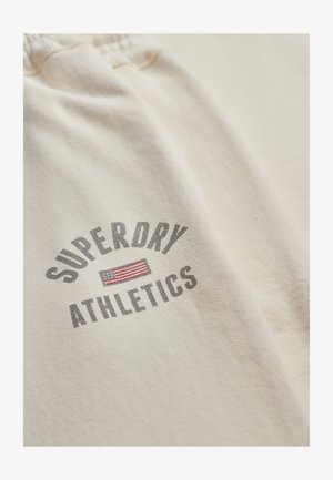 Beżowe spodnie dresowe Superdry Athletics z elastycznym pasem i nadrukowanym logo, które przedstawia mały prostokąt w czerwono-białe paski.
