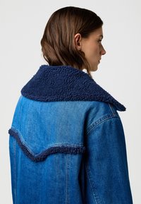 Kurtka dżinsowa z granatowym, puszystym kołnierzem, o strukturalnym kroju i kontrastujących, niebieskich fakturach. Charakterystyczne detale na tylnej części pachy.