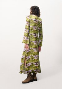 Langes grünes Kleid mit einem Landschaftsmuster in lila und rosa Tönen, mit einem tiefen Schlitz an der Seite und Glockenärmeln, kombiniert mit braunen Stiefeln.