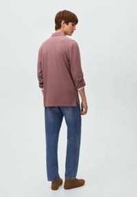 Giovane uomo in piedi di lato, indossa una camicia a maniche lunghe color malva, jeans blu e stivali alla caviglia marroni, su uno sfondo chiaro e uniforme.