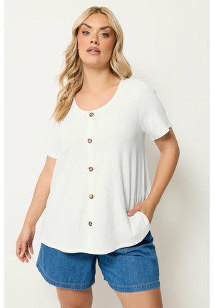 Kvinde iført en hvid kortærmet knapbluse og blå cowboyshorts, stående med den ene hånd i lommen mod en ensfarvet baggrund.