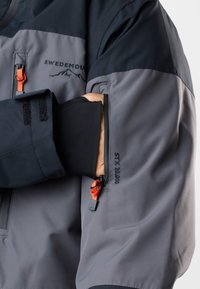 Personne portant une veste grise et noire avec des tirettes de fermeture éclair orange, la main partiellement dans une poche zippée au bras étiquetée "STX 20 000".