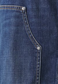 Denimjakke med en dyp blå farge, kontraststing og metalliske detaljer på frontlommen. Teksturert stoff synlig.