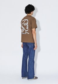 T-shirt marron à manches courtes avec un contour blanc d'un visage au dos. Assorti à un jean en denim évasé bleu et des chaussures foncées.