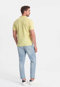 Lys gul polo skjorte med korte ærmer, kombineret med lyseblå jeans, set fra bagvyn. Har krave og sideskår. Hvide sneakers.