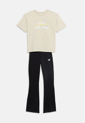 T-shirt beige New Balance con motivo floreale giallo abbinata a pantaloni neri svasati con logo New Balance sulla coscia sinistra.